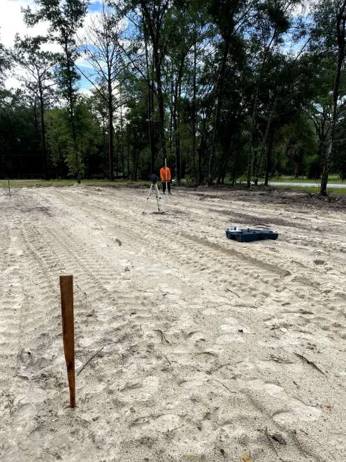 Wooded dirt clearing with a wooden stake in the foreground and a person in orange in the distance
