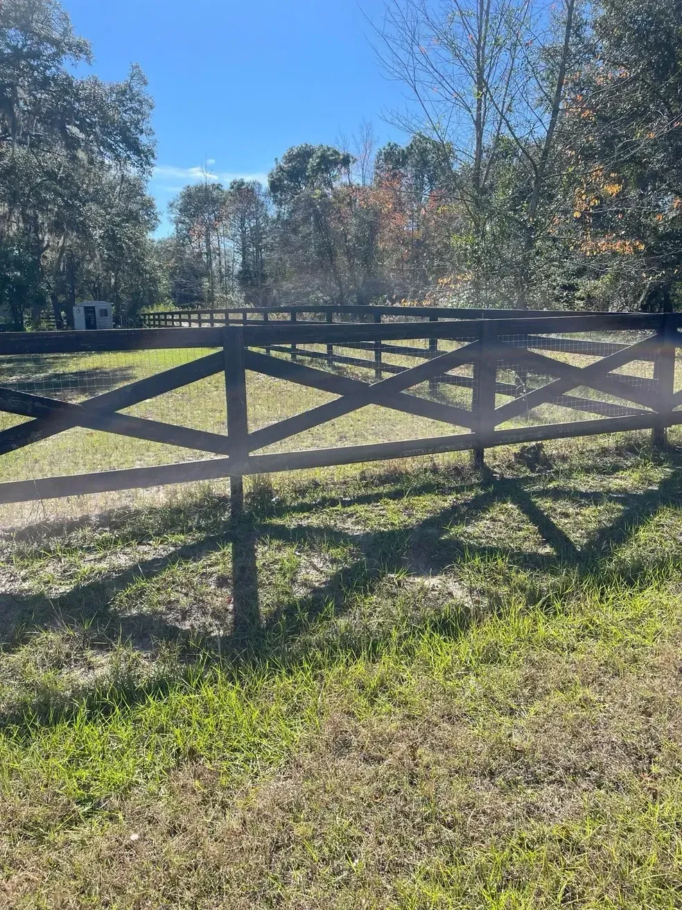 Wooden fence beside a grassy field under clear blue sky, with trees in the background.