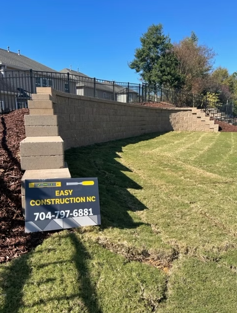 Retaining wall and steps made of tan blocks in a backyard, with a sign for Easy Construction NC.