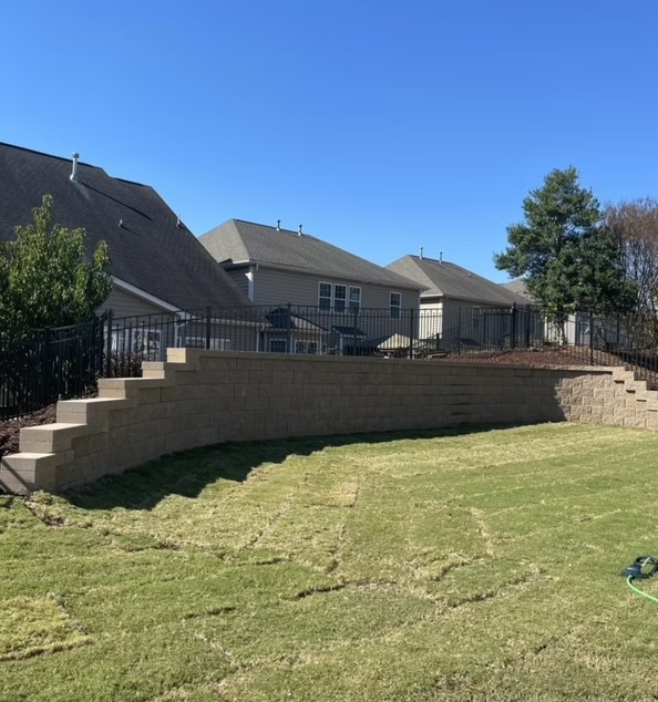 Backyard with retaining wall and steps, grass, houses, and clear sky.