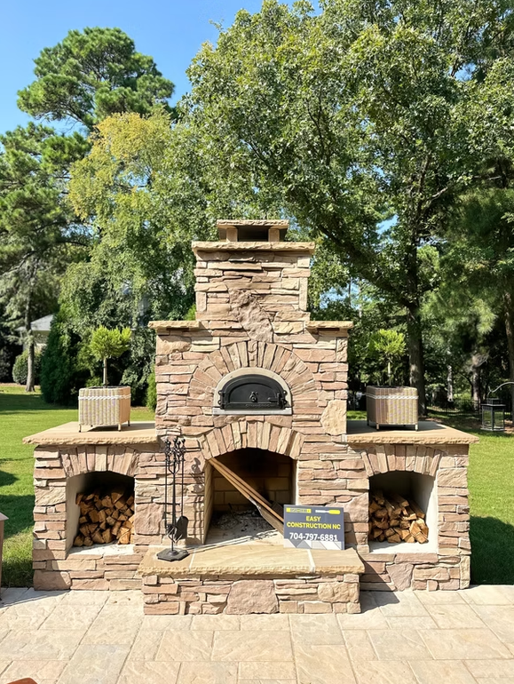 Stone outdoor pizza oven with stacked wood, set on a patio in a sunny yard.