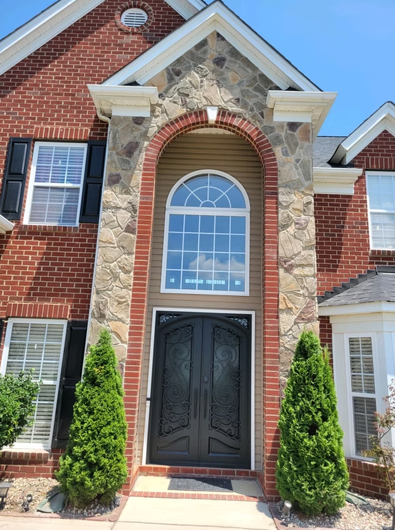 Red brick house with stone archway over black double doors, two green bushes flank the entrance.