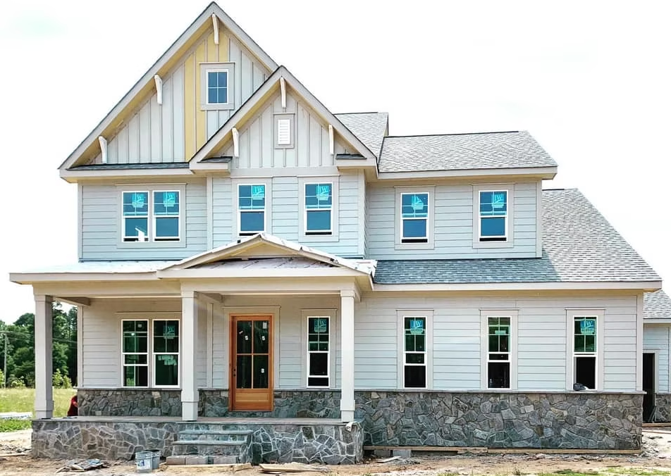 Two-story light blue house with stone foundation, porch, and unfinished construction site.