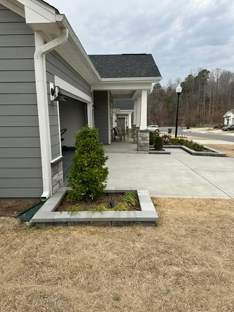 Gray house exterior with a garage and covered porch, concrete walkway, and a small tree in a raised garden bed.