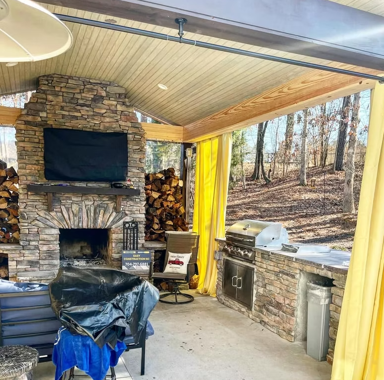 Outdoor living space with stone fireplace, TV, grill, and yellow curtains.