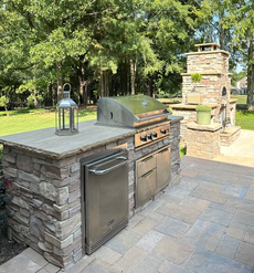 Outdoor stone fireplace with TV, grill, yellow curtains, and wooded view.