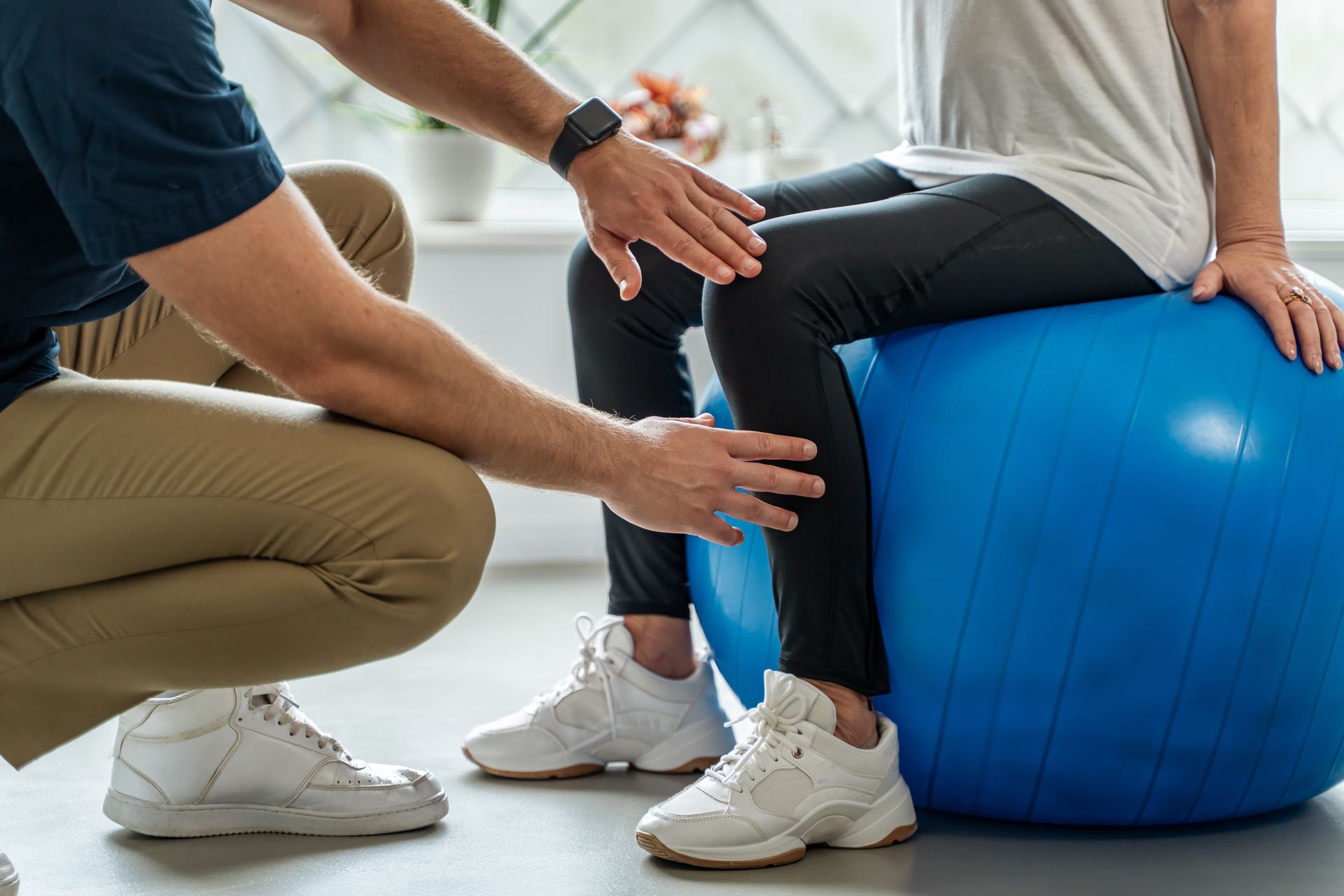 Therapist assisting a person sitting on exercise ball, touching the knee, in a brightly lit room.