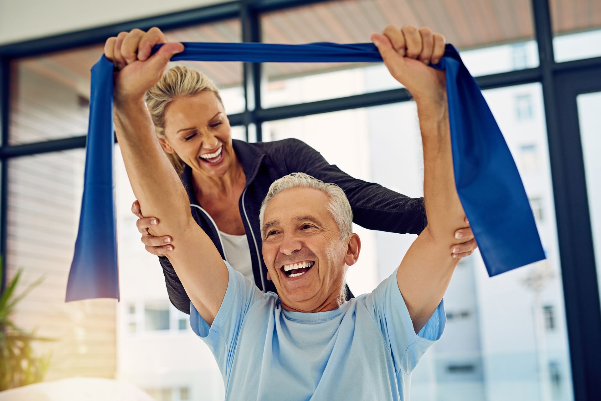 Woman assists man with stretching resistance band, both smiling. Blue band, bright room.