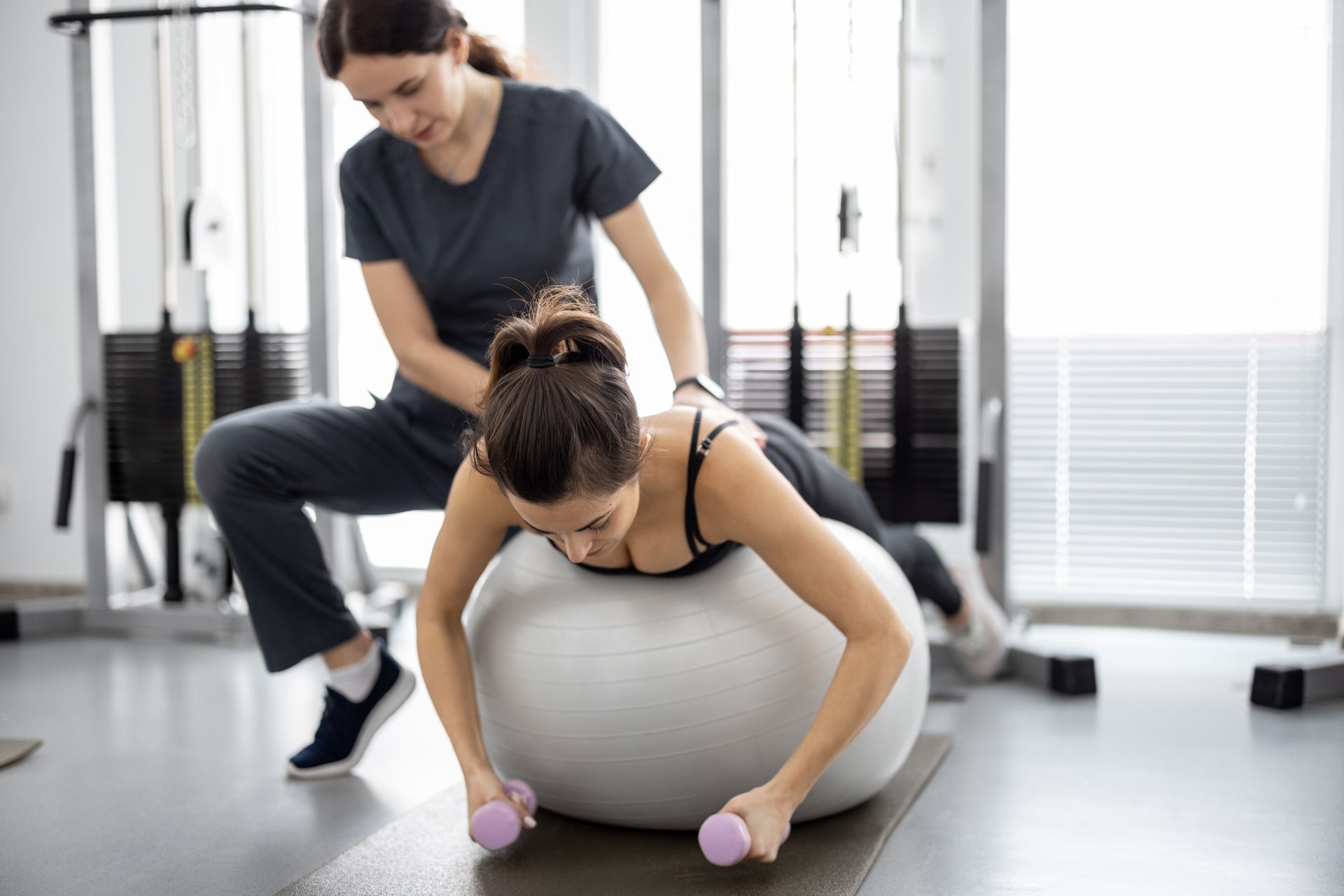Woman exercising on exercise ball with assistance from a therapist in a gym.