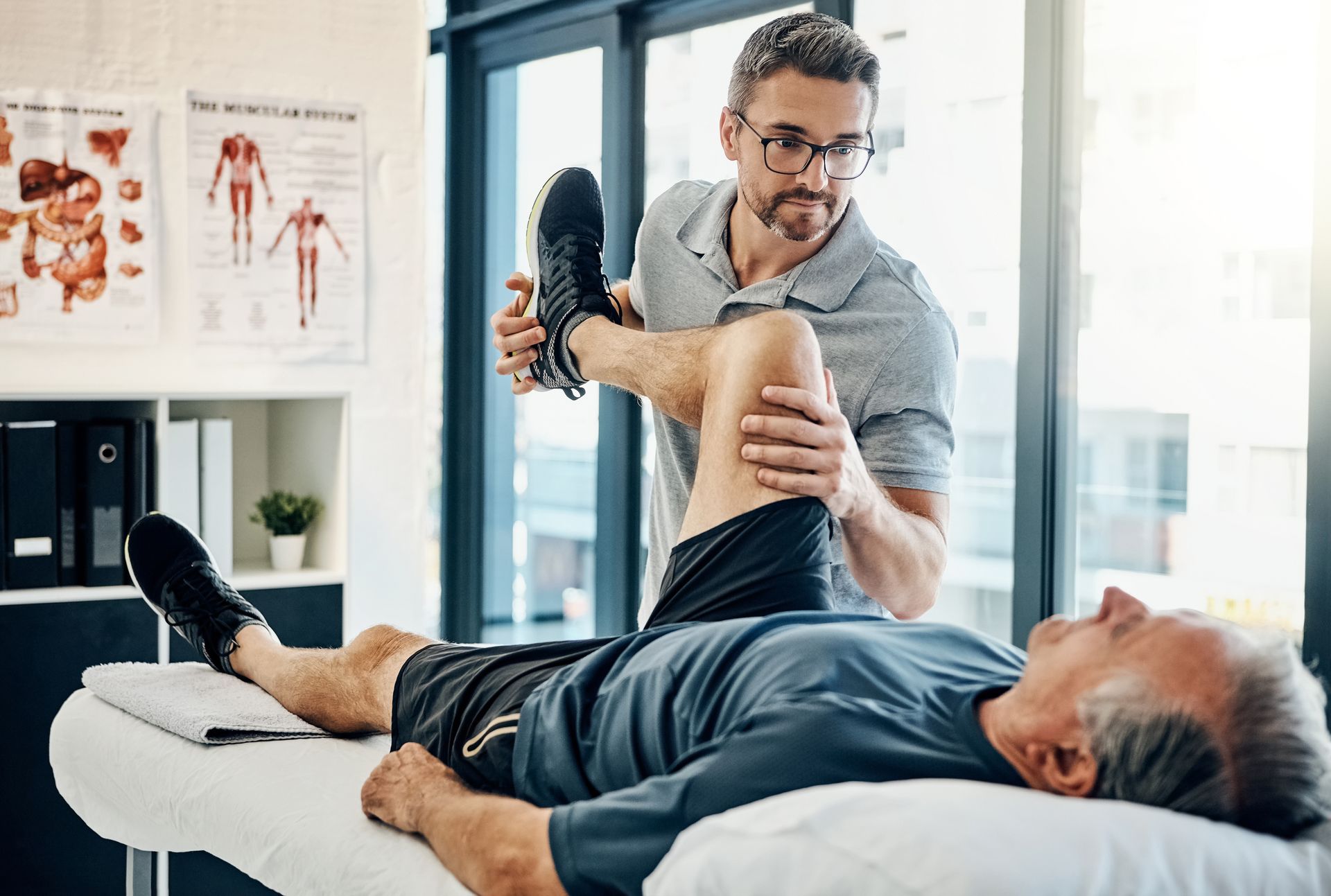 Physical therapist stretching patient's leg on exam table. Room with medical posters and window.