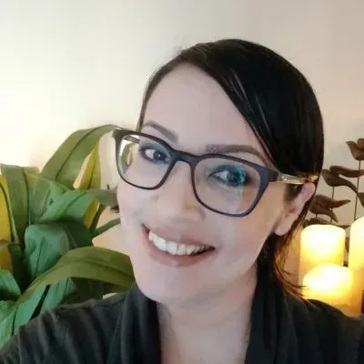 A woman wearing glasses is smiling in front of a plant and candles.