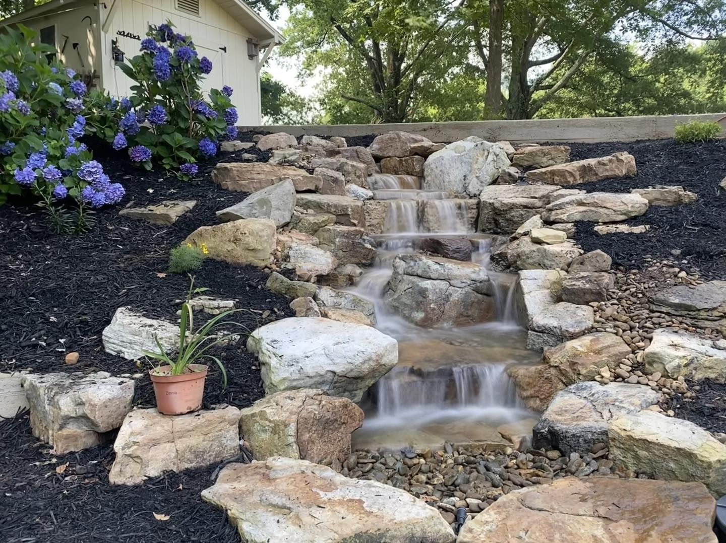 Stone garden waterfall with flowing water, purple flowers, and potted plants beside a house
