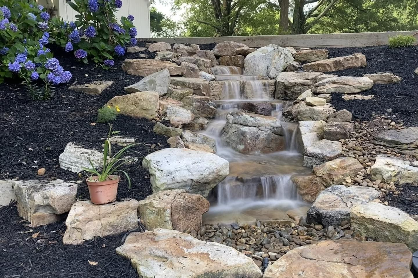Rock garden waterfall with cascading water over stones and a purple flower bush nearby