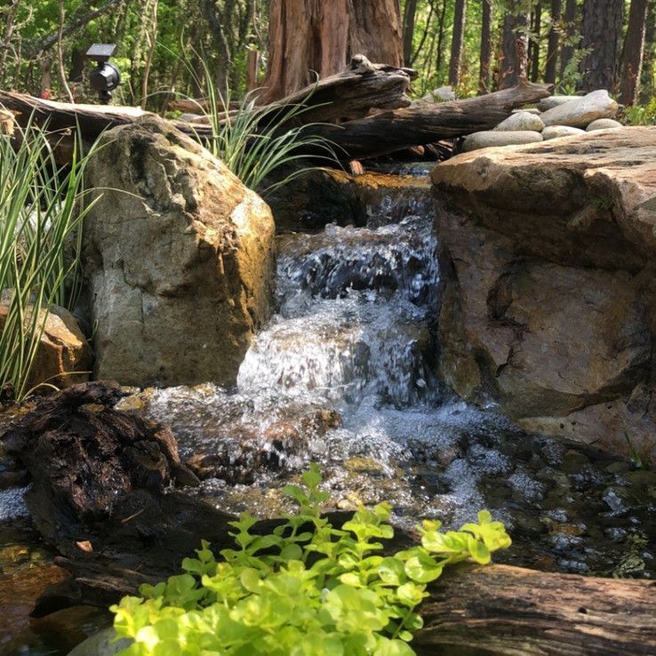 Small waterfall flowing over rocks in a garden, with green plants in the foreground.