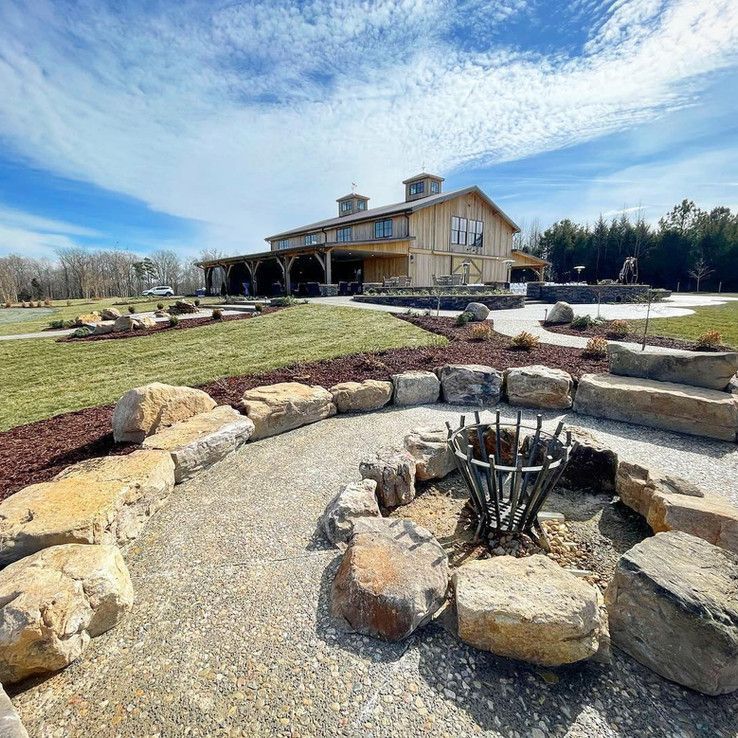 Stone fire pit in a landscaped yard with a rustic barn-style house and blue sky in the background