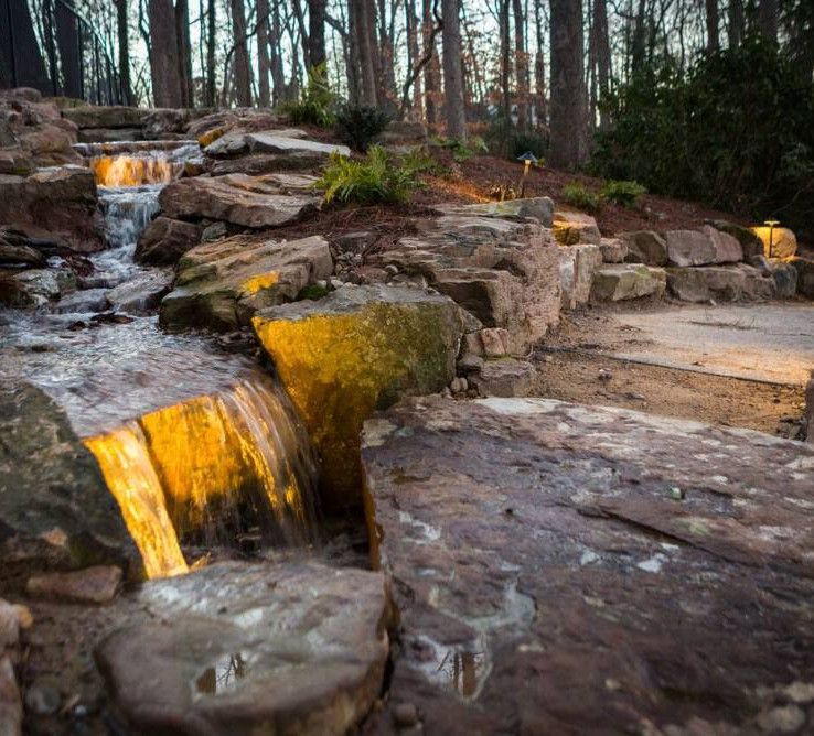 Stone waterfall cascading into a rocky forest stream at dusk, with warm golden light on the water