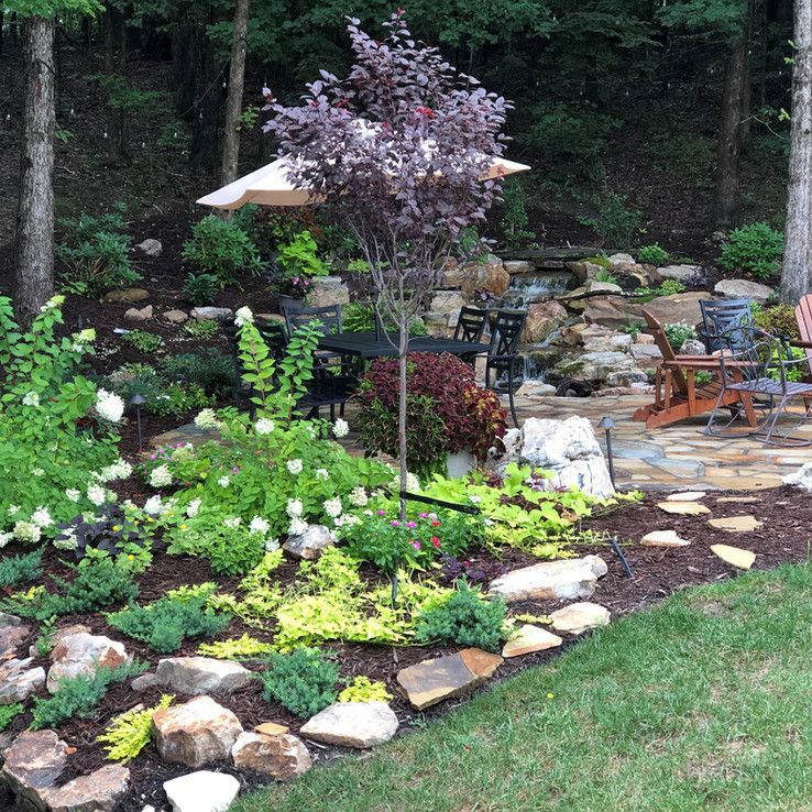 Lush backyard garden with stone border, flowering plants, patio chairs, and a shaded umbrella near a wooded edge