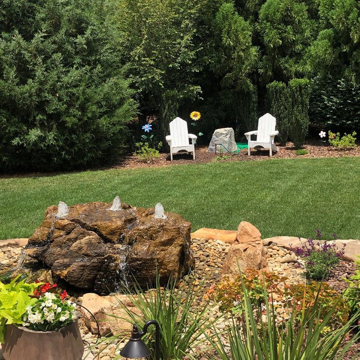 Backyard garden with rocks, flowers, white chairs, and a large tree line
