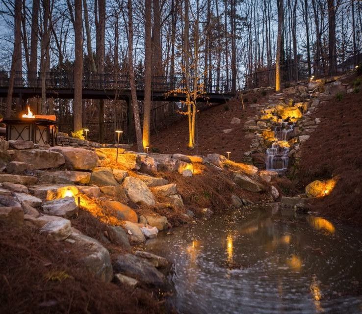 Lit pond waterfall with glowing landscape lights among rocks and bare trees at dusk