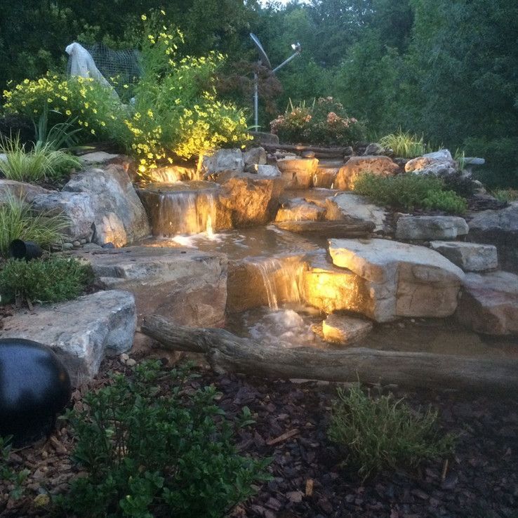 Illuminated backyard waterfall cascading over stacked rocks at dusk, surrounded by lush greenery.