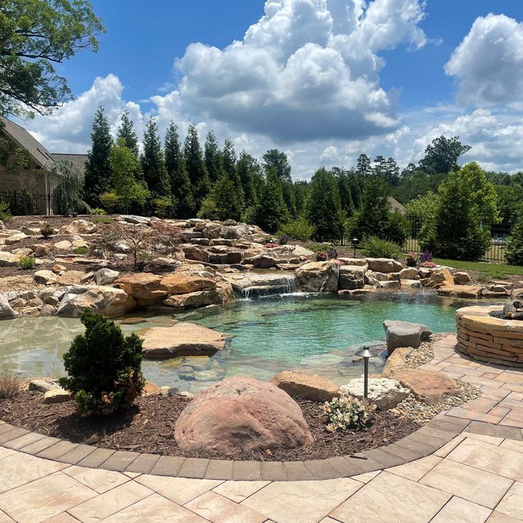 Rocky backyard pool with clear turquoise water, stone patio, and tall trees under a cloudy blue sky