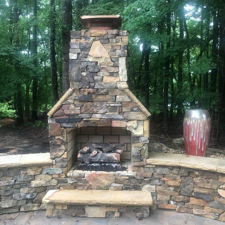 Stone outdoor fireplace with bench seats in a wooded park, next to a red ceramic planter