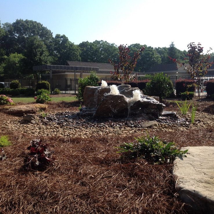 Landscaped garden with a rock fountain, mulch beds, shrubs, and a building in the background.