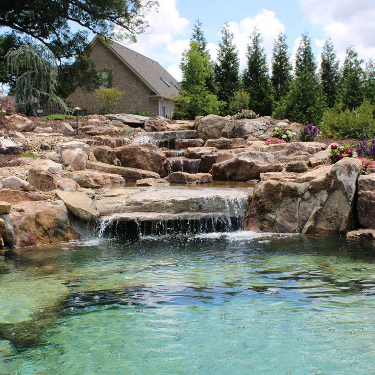 Rock waterfall cascading into a turquoise pond beside a landscaped garden and house