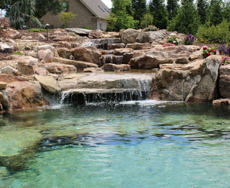 Rock waterfall cascading into a clear turquoise backyard pond with rocks and greenery around it