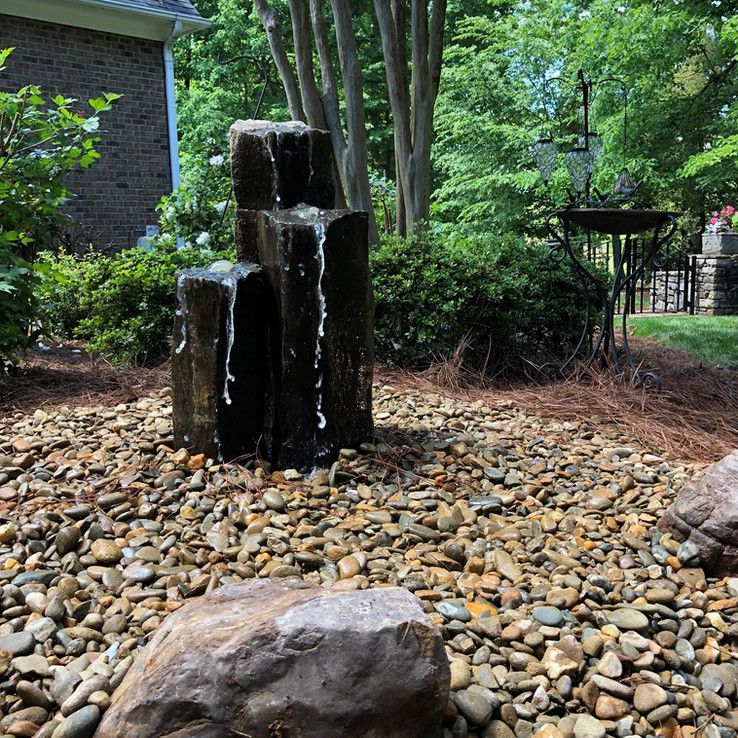 Stacked stone fountain in a landscaped garden, water cascading over dark columns onto river rocks.