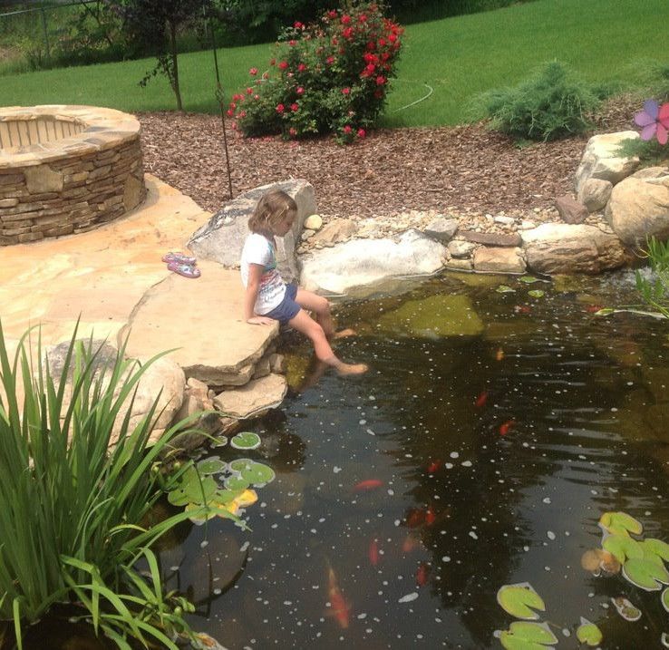 Child sitting by a garden pond, dangling feet in the water beside rocks and flowers