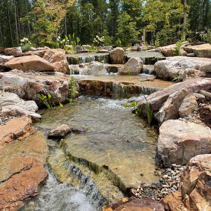Rock garden stream with cascading water over stone steps, surrounded by trees and boulders.