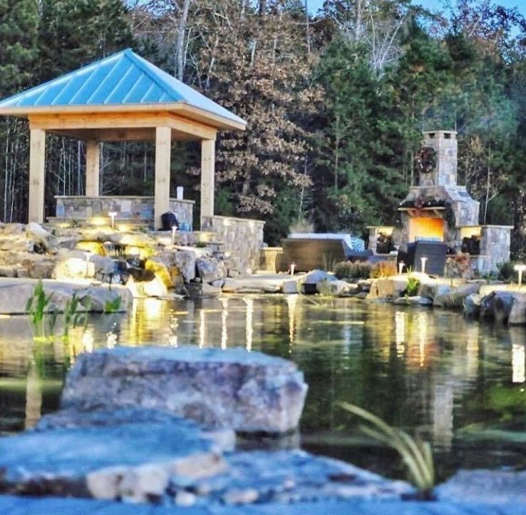Scenic pond with stone waterfall, lit pavilion, and trees reflected in calm water