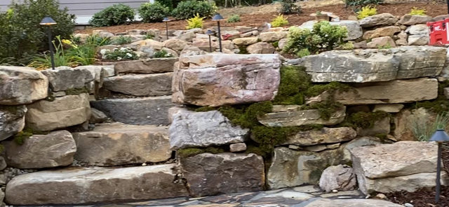 Stone retaining wall with stacked rocks and small plants on top, in an outdoor garden setting
