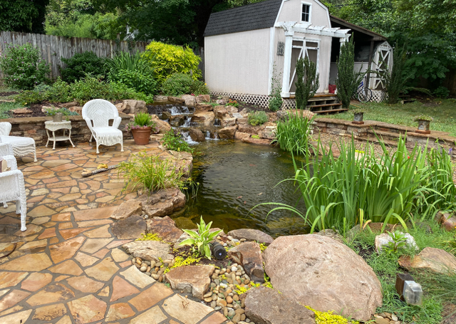 Backyard pond garden with stone patio, white chairs, and a small shed amidst greenery