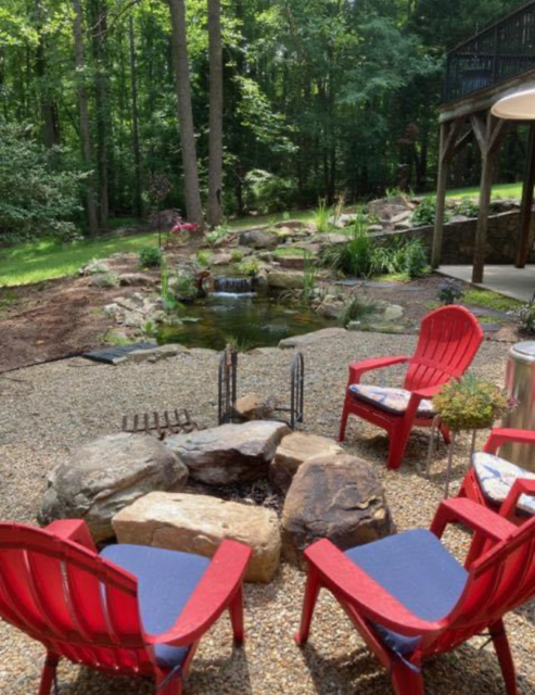Red Adirondack chairs around a fire pit beside a wooded backyard stream and garden path.
