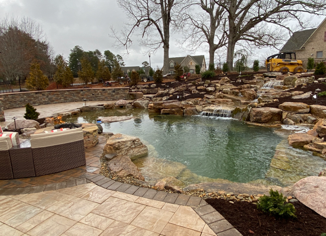 Stone-lined backyard pool with waterfall, hot tub, and bare trees in a landscaped patio area