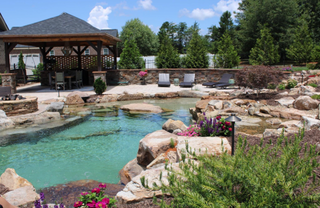 Backyard pool with turquoise water, rock waterfall, gazebo, and colorful flowers on a sunny day