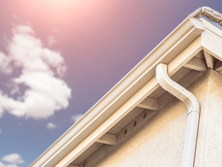 Beige house gutter and downspout against a sky with clouds.