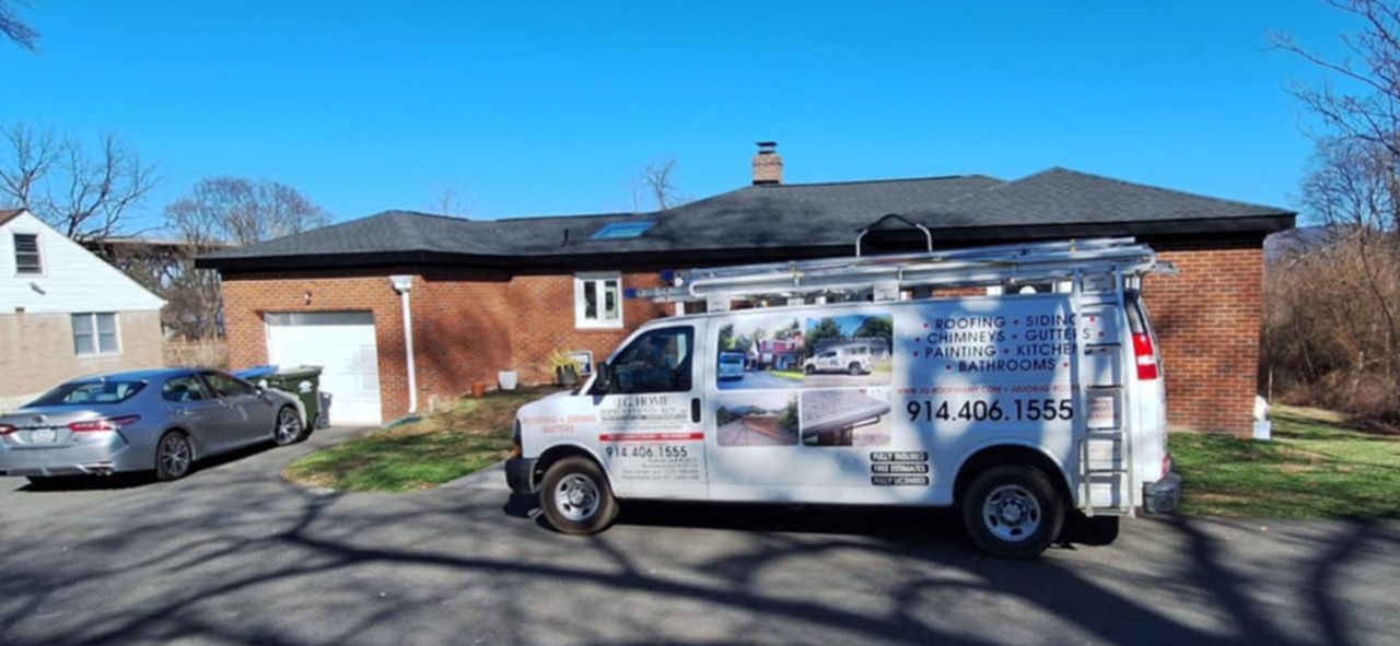 A white contractor van with ladder parked in front of a brick house with a gray car in the driveway on a sunny day.