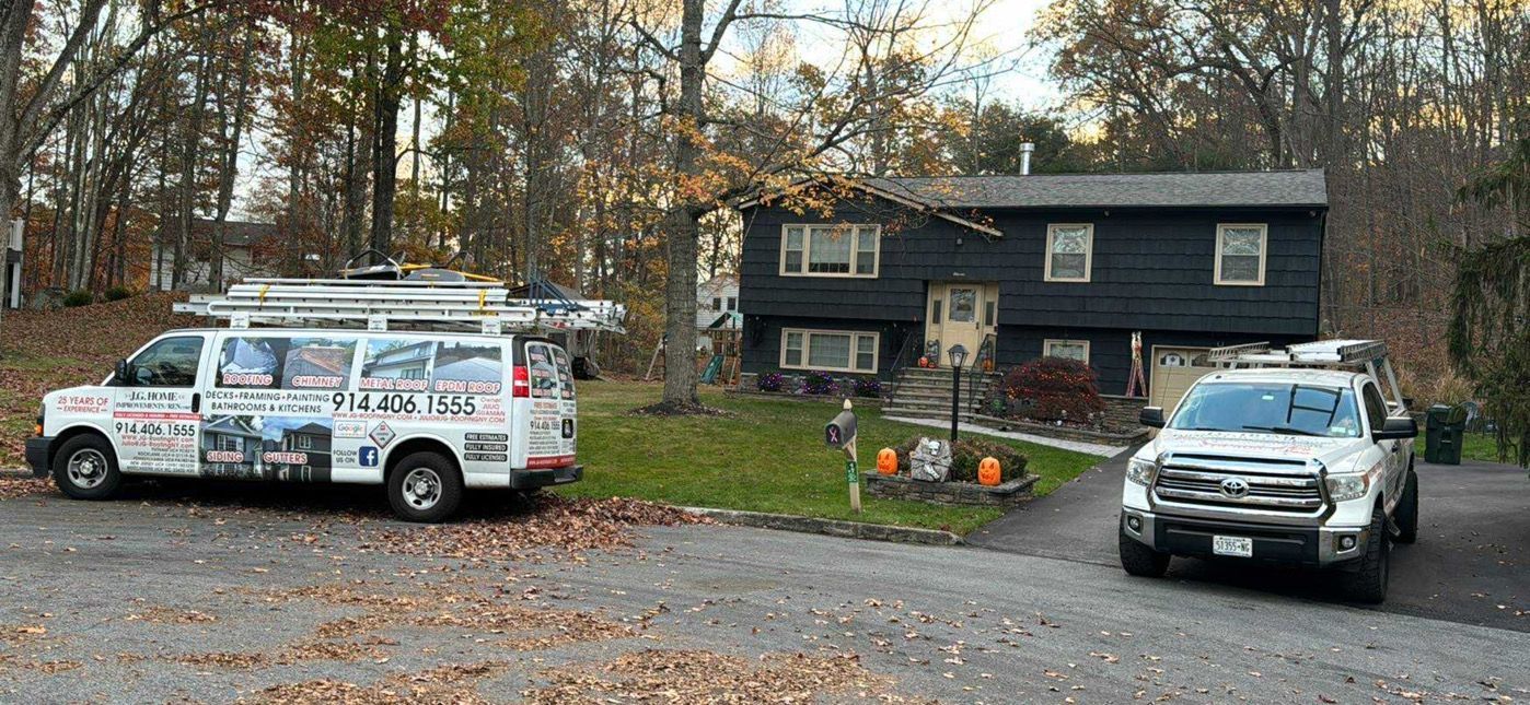 White van and pickup truck parked in front of a dark-colored house. Trees in the background, leaves on the ground.
