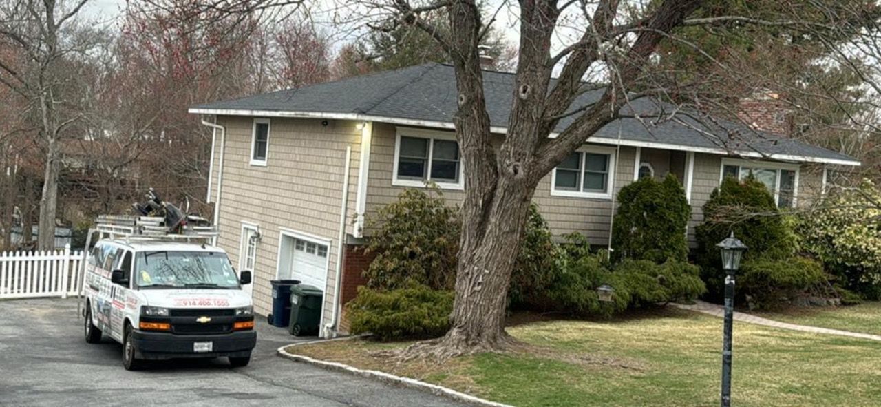 A two-story beige house with a black roof, a tree, and a white van parked in front.
