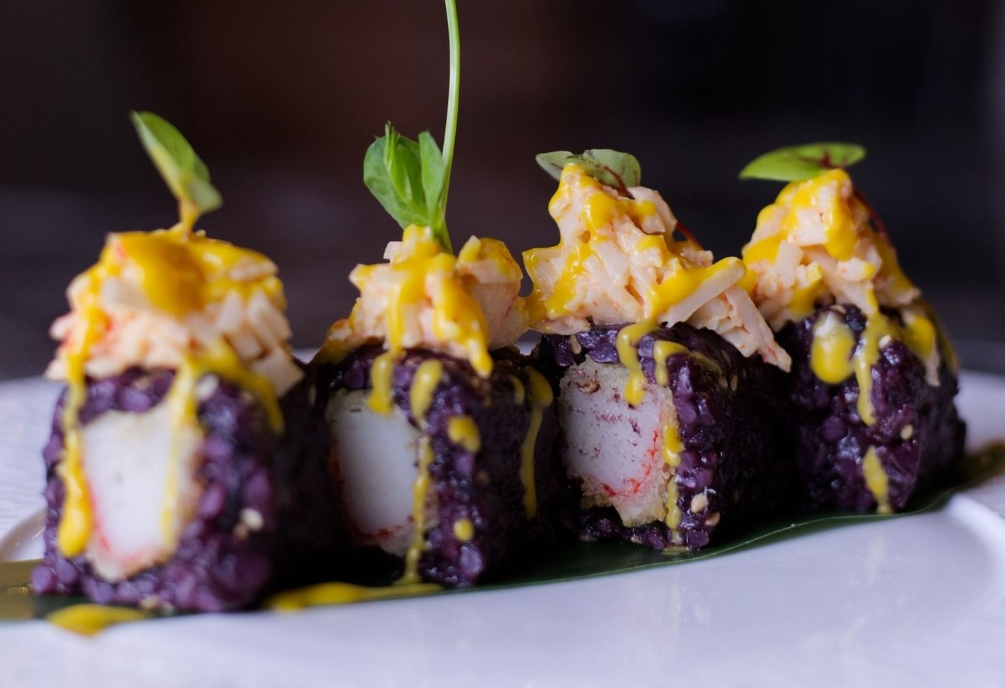 Quattro pezzi di sushi preparati con riso viola e granchio, conditi con salsa gialla e microverdure su una foglia di banana.
