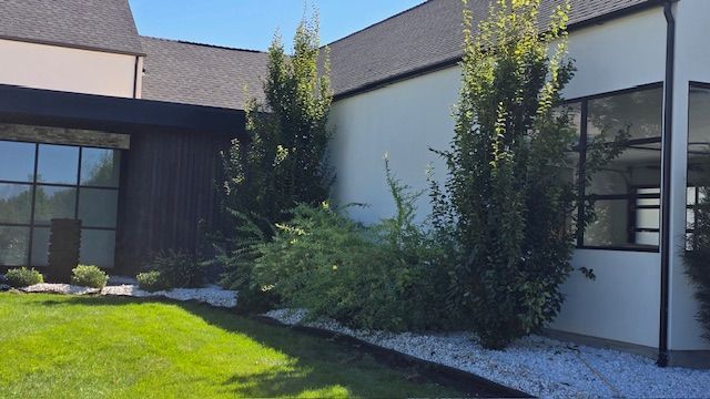Exterior of a building with dark trim and windows, light-colored walls, and green trees and grass with a white rock bed.