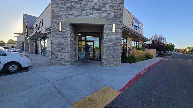 Exterior of a stone-faced building with awnings. A person stands at the entrance. Sidewalk and parking area in view.