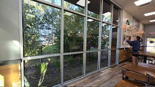 Inside a building, a large window reveals trees and a person cleaning.