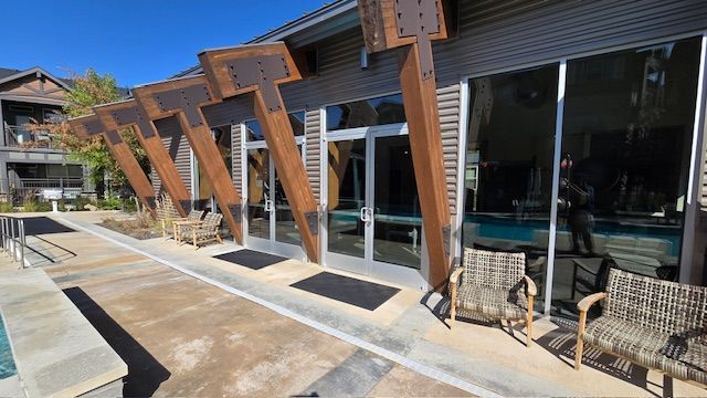 Poolside building with large glass windows and geometric wooden beams. Two chairs sit outside the entrance.