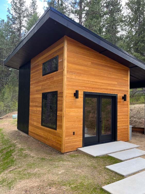 Modern cabin with wood siding, black accents, and a sloping roof. Concrete path leads to double doors.