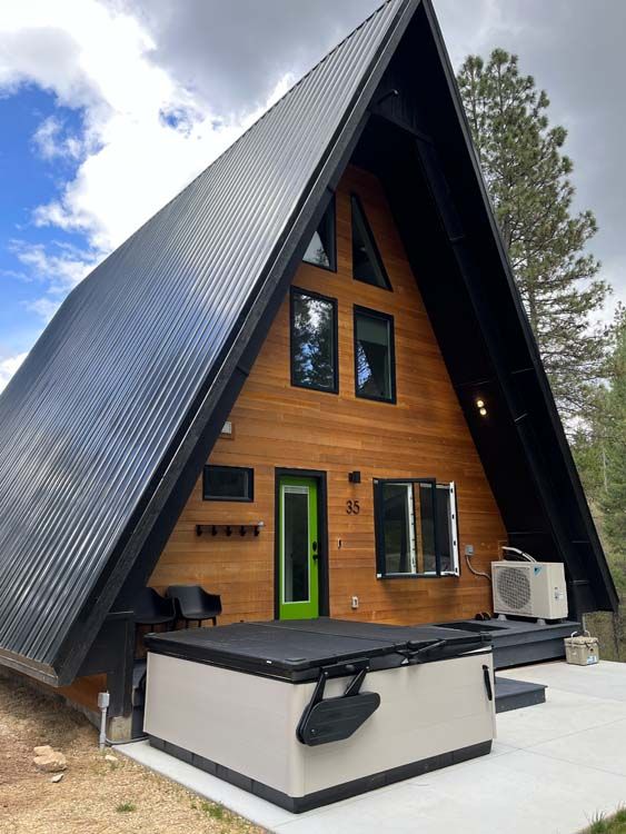 A-frame cabin with black roof, wooden siding, and hot tub. Green door and windows.