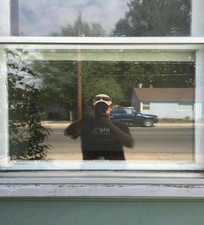 Reflection in window shows person holding phone, wearing a hat and shirt, with a street and houses in background.